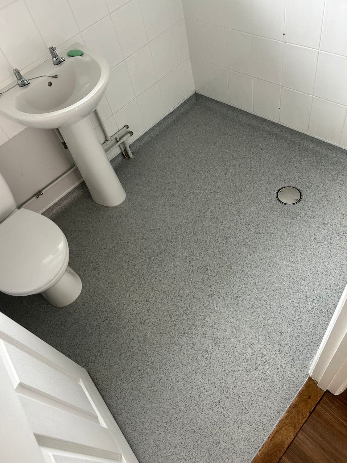 Grey speckled wet room flooring coved up to white tiled walls, with a sealed floor drain beside a pedestal basin and toilet