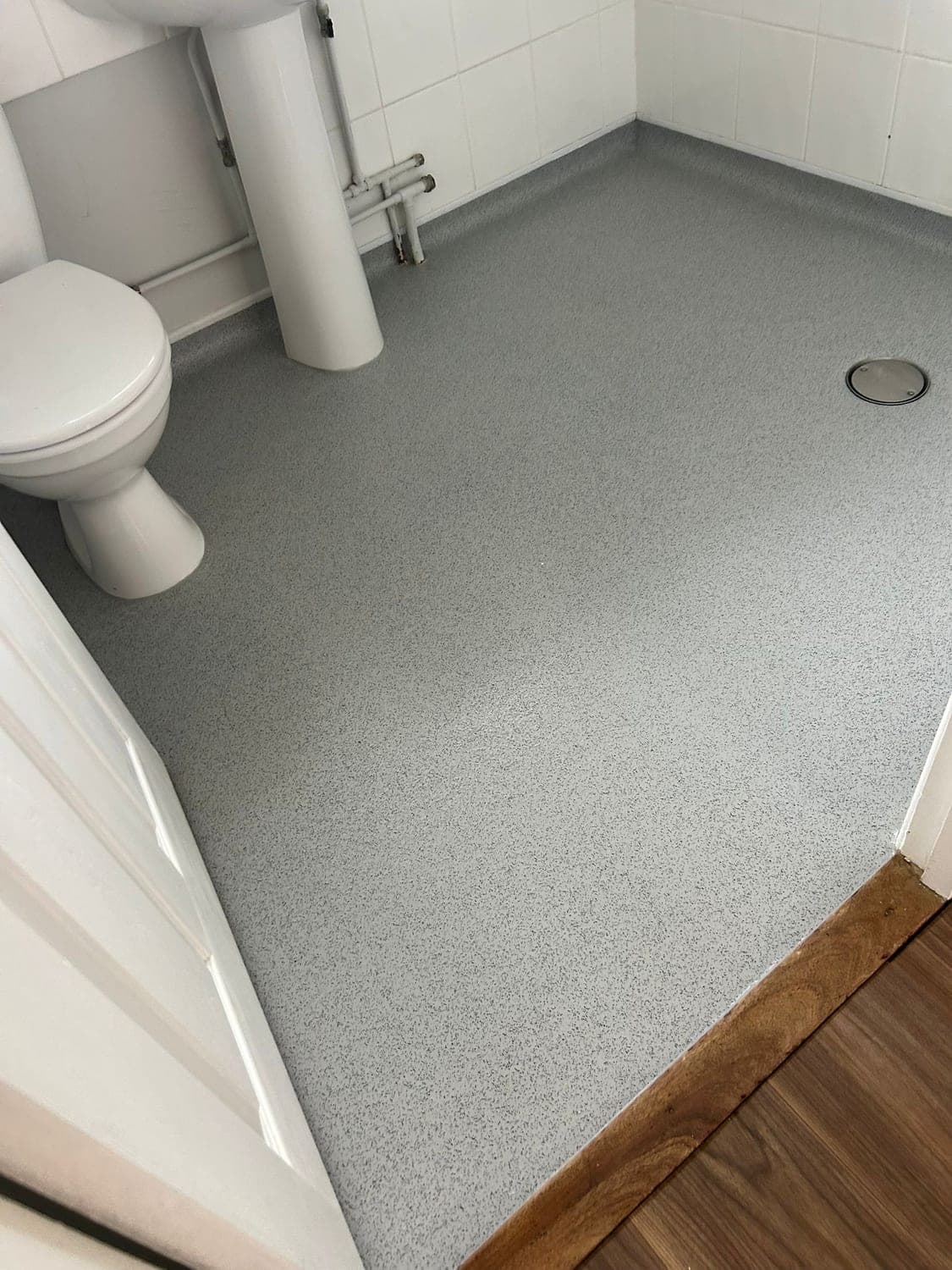 Slip-resistant grey wet room floor coved up the walls and finished flush at the doorway against wood-effect flooring, with no threshold to step over